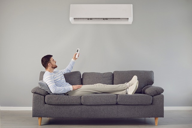 A guy sits stretched on a sofa holding a remote control aimed at an air conditioning unit