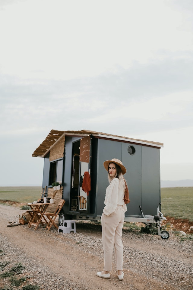 A woman stands  on the side of a tiny house next to a gravel road