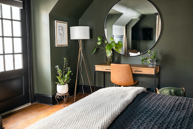 A furnished bedroom with a large round mirror on the wall, a simple desk, and chair on the side, and a few potted plants