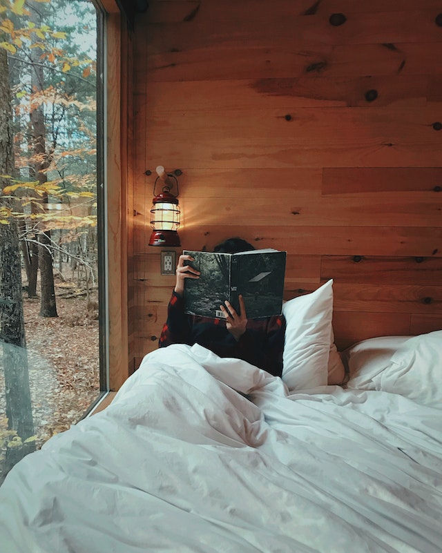 Person reads a book on the bed right next to a wide glass window